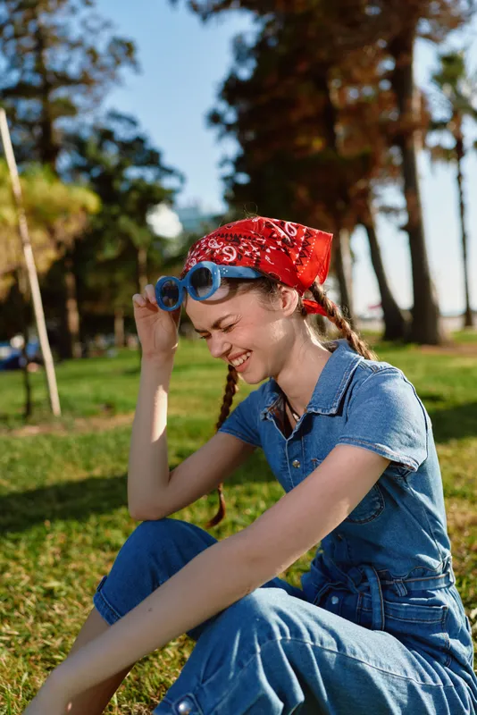 lifestyle woman in modern boho-western denim street style wearing a red bandana and blue sunglasses, smiling outdoors with natural film color tones, casual summer vibe
