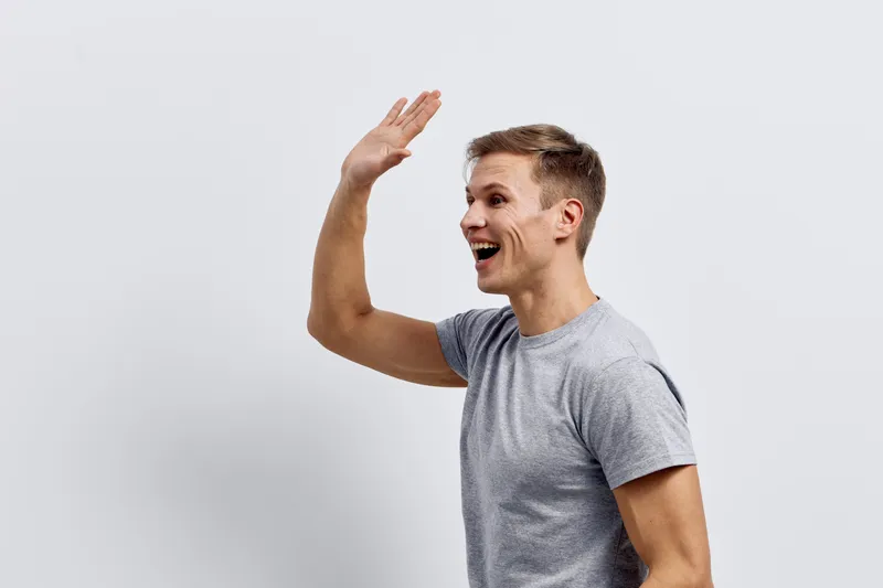 Smiling man waving hand in greeting, casual gray t-shirt, happy expression, friendly gesture, indoors, neutral background, lifestyle concept, positive mood, young adult male, cheerful attitude, relaxe