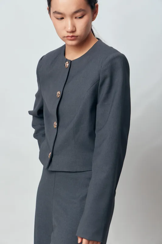 Young woman wearing gray suit with buttoned jacket and matching pants standing with downcast eyes against plain background. Fashion portrait with minimalistic style and neutral colors.