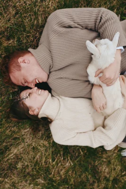 Couple lies on grassy ground outdoors, embracing in cozy sweaters, holding a gentle goat kid, warm romance and tenderness, peaceful countryside mood A lifestyle moment of affection now