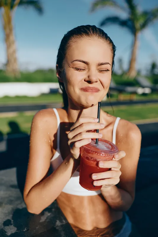 Bright outdoor scene with a woman sipping a pink smoothie, eyes closed, sunlit smile, poolside breeze, casual attire, vibrant colors, warm summer mood, refreshing moment in nature