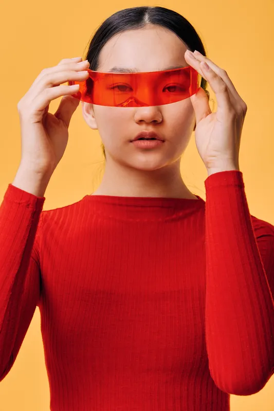 Young woman wearing futuristic red sunglasses against orange background. Stylish female model with straight hair and red sweater posing indoors with high fashion eyewear accessories.