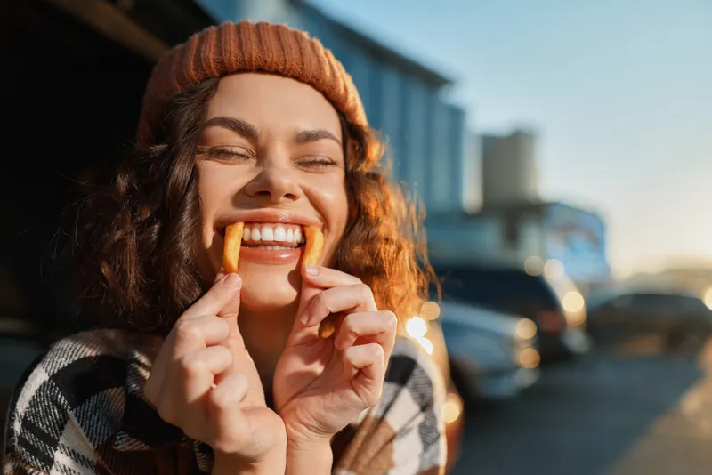 Woman smile with fries held as playful mustache at golden hour, candid lifestyle portrait in urban street, beanie and snack joy conveying authenticity, mindful living and emotional storytelling.