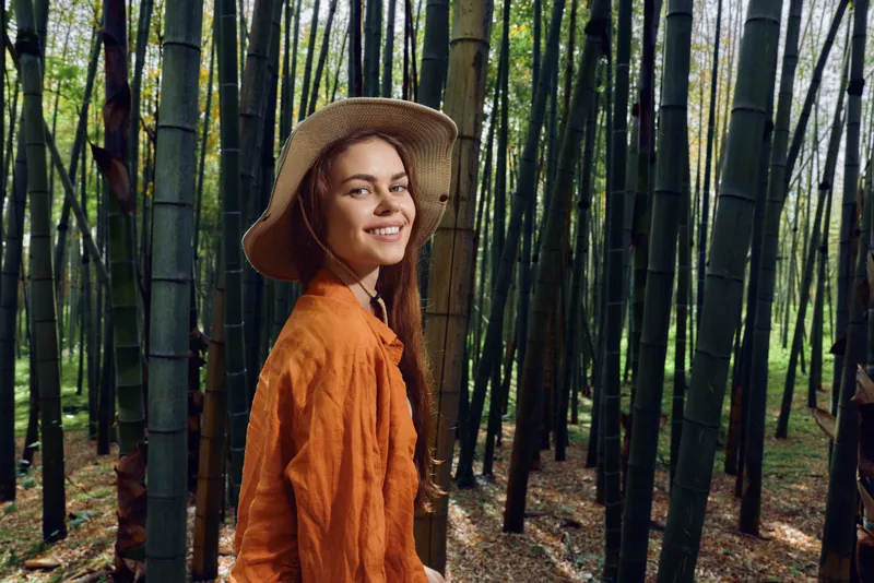 Woman in straw hat smiles in a bamboo forest portrait, wearing orange shirt during outdoor nature walk. Travel vibe, relaxed expression and green stalks creating a serene background.