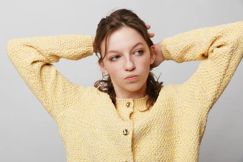 Young woman in yellow knitted sweater stretches with hands behind head, looking sideways. Casual style, relaxed mood, isolated on light grey background. Concept of comfort and leisure.