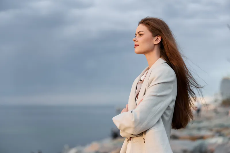 Woman standing in thoughtful pose by the sea wearing beige jacket with long hair and cloudy sky background in calm natural light