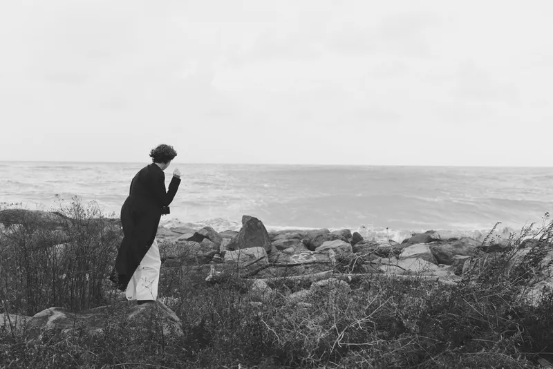 Monochrome scene of a solitary figure on a rocky coast, gazing toward the sea, wind in hair, contemplative mood, rugged shoreline, minimal and dramatic.