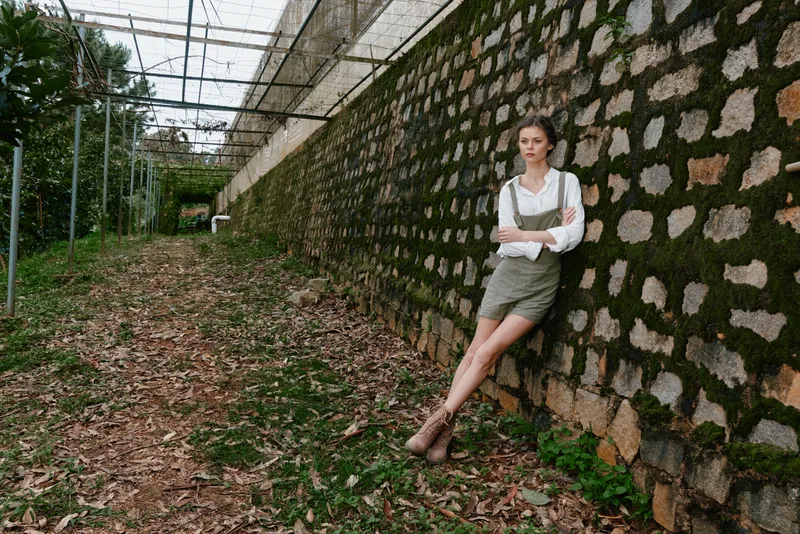 Young woman in casual summer outfit leaning against a moss covered wall, showcasing emotions of confidence and tranquility in a natural outdoor setting