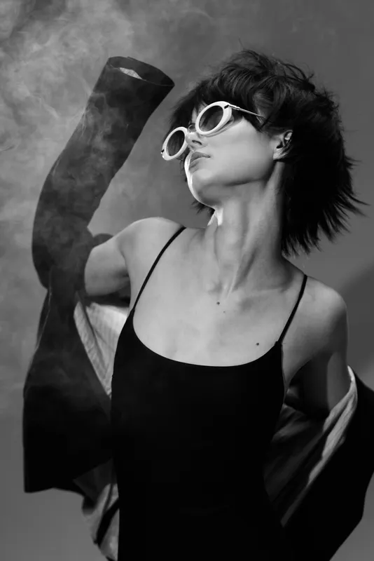 Black and white portrait of a stylish woman wearing sunglasses and a dress with flowing fabric in smoky background.