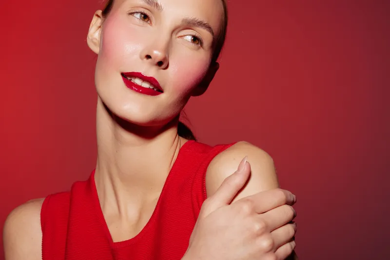 Young woman with red lipstick and sleeveless top poses against red background. Concept of beauty, fashion, and confident female portrait with vibrant color contrast.