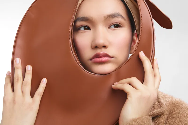 asian woman wears a large brown mask in a studio setting, creating a playful and mysterious mood as she gazes softly through the opening.