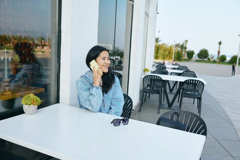 Smiling woman on outdoor cafe talks on phone beside a white table, casual seating, glass storefront, sunglasses on surface, sunlight highlights relaxed urban conversation in a modern setting