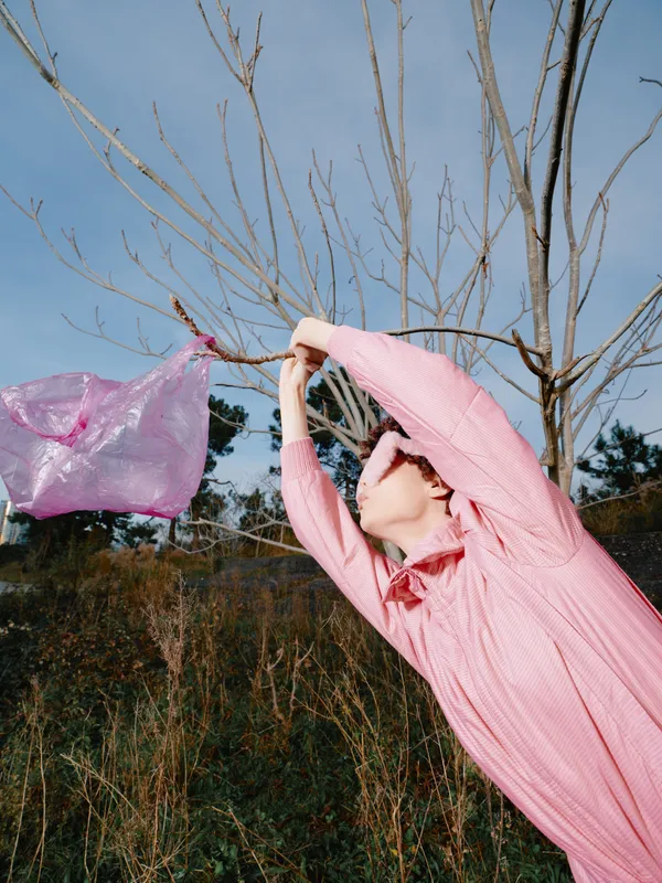 Woman in a pink dress swings fabric from a bare tree in a rural field, wind catching the cloth, creating motion and a sense of freedom.