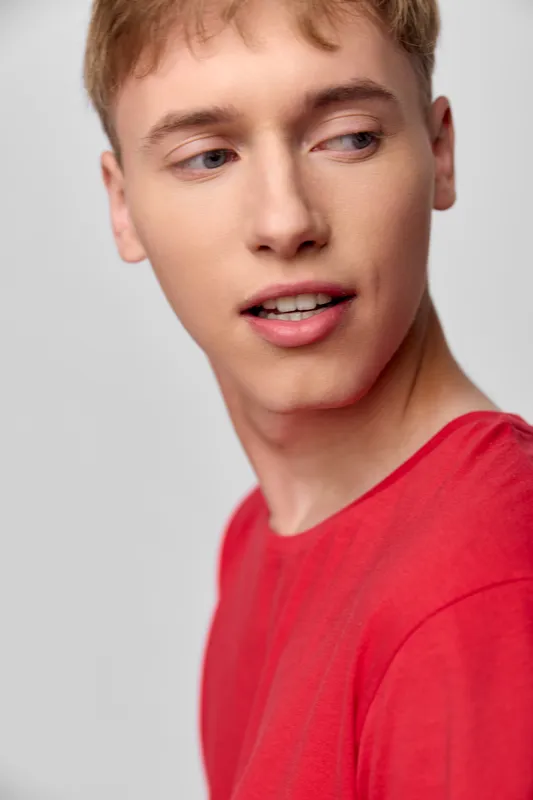 Young man in a red shirt glances sideways with a thoughtful expression, closeup of face and casual style against a clean background, suitable for fashion and lifestyle in stock imagery for marketing