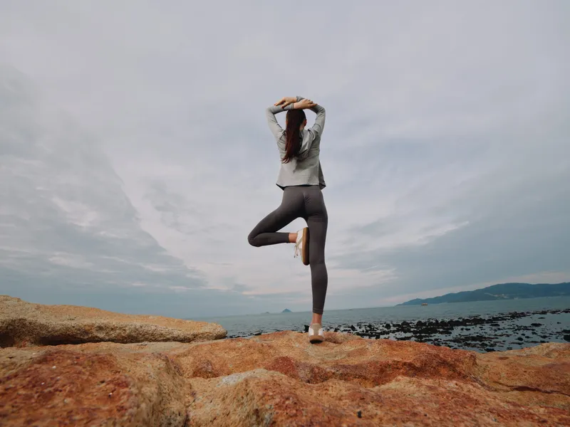 Young woman practicing yoga on rocky beach, promoting mindfulness and wellness through nature, wearing comfortable athletic wear, sunset atmosphere
