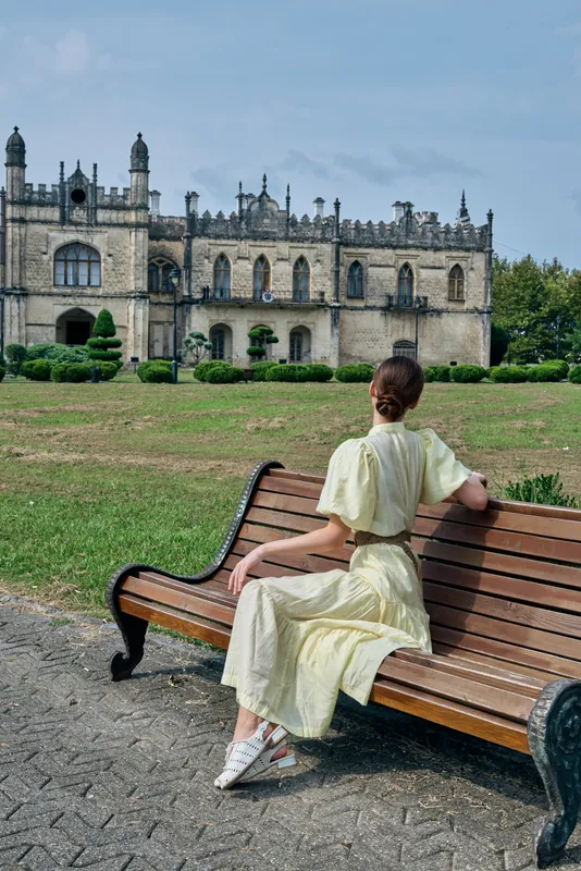 A person sits on a wooden bench in a well kept garden, wearing a light dress, facing a grand castle with ornate architecture.
