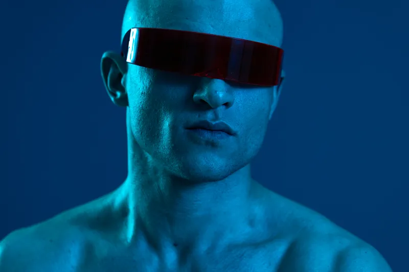 Confident man wearing red sunglasses, showcasing a unique look in a blue light setting. The image evokes intrigue with strong visual elements and bold contrasts.