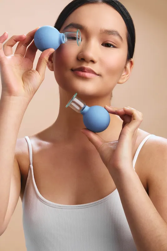 Asian woman is seen holding medical bulb syringes near her face in a bright studio. She wears a white tank top and projects calm confidence, creating a clean, clinical lifestyle vibe.