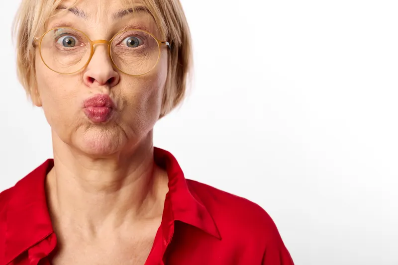 Surprised woman with wide eyes in a red shirt closeup, puckered lips and expression, wearing glasses, studio portrait on white background, showing strong surprise and shock emotion