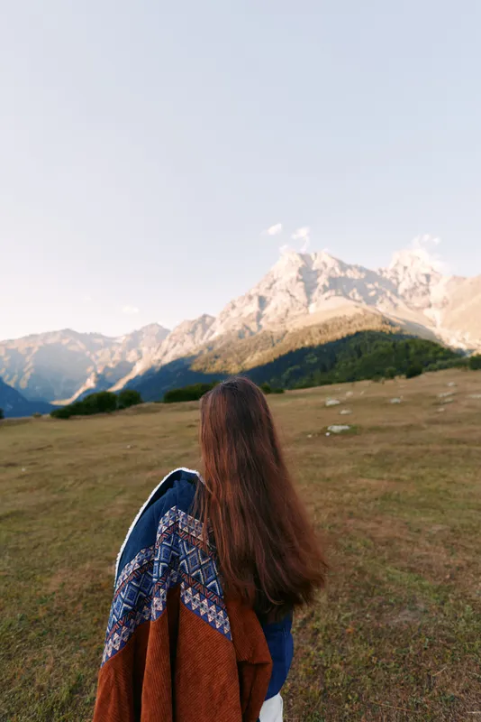 Woman back with longhair and shawl standing in meadow facing mountains, landscape and nature at golden hour. Solo traveler, peaceful outdoor scene with hiking and panoramic vista.