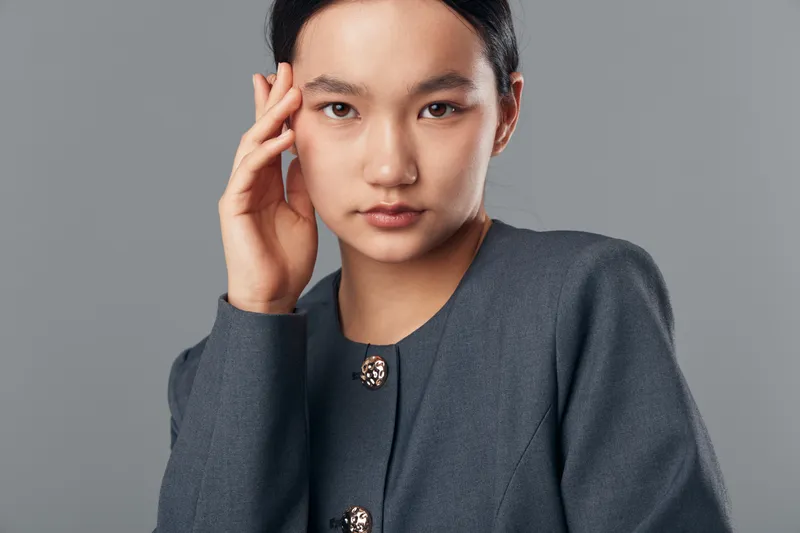 Young Asian woman in gray jacket with clasp buttons posing with hand near head on gray background. Closeup portrait with serious expression for fashion or business concept.