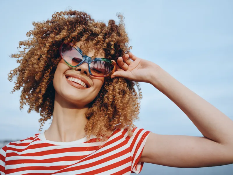 Bright portrait of a joyful woman with curly hair wearing colorful sunglasses and a red striped shirt, smiling against a clear blue sky.