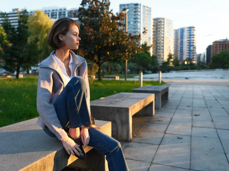 A thoughtful professional woman sits on a concrete bench in an urban park, wearing jeans and a light jacket. She gazes into the distance, sunlight warming the scene and conveying calm focus amid a
