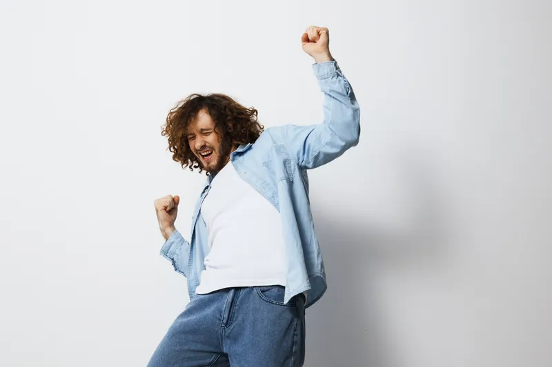 Joyful man celebrating success with vibrant energy in casual attire, showcasing curly hair against a minimalistic white background