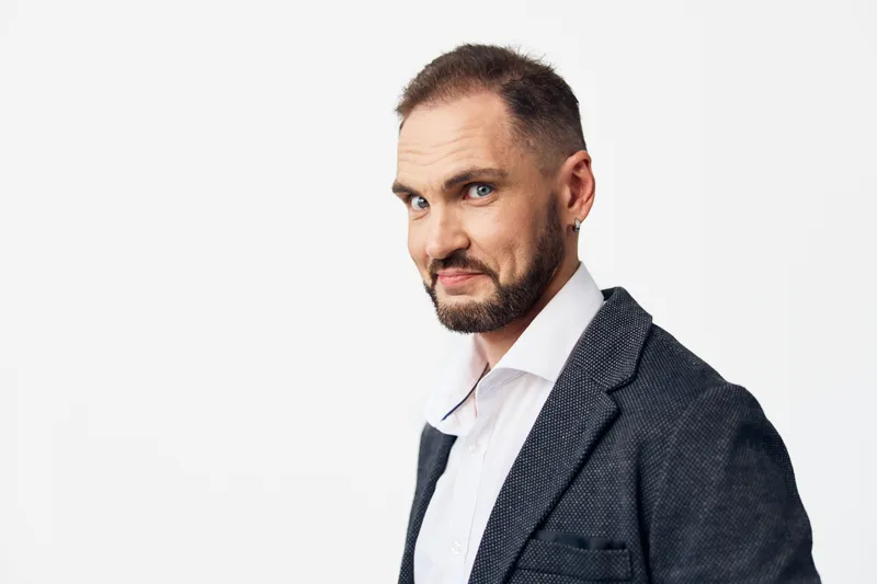 A focused business man stands against a plain colored backdrop, conveying confidence and calm emotion, refined style, and professional presence in a modern work setting.