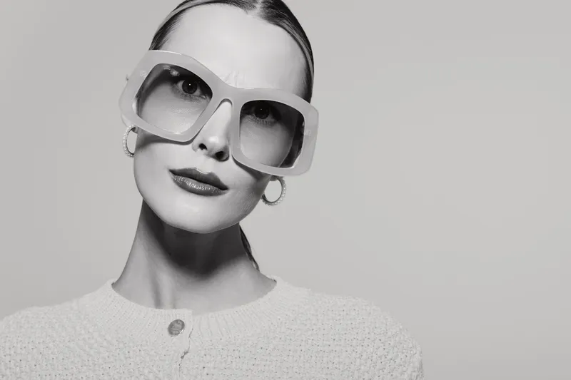 Portrait of a fair skinned Caucasian woman wearing large sunglasses and hoop earrings, dressed in a knitted sweater, looking confidently at the camera in a studio setting.
