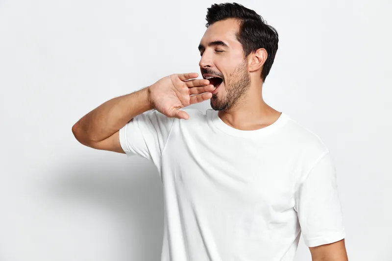 Tired young man yawning with hand over mouth, wearing a plain white t-shirt, isolated on solid light grey background. Emotion and daily life concept.