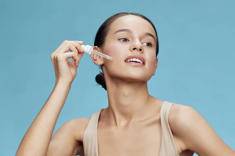 Young woman applying skincare serum with dropper, showcasing healthy skin, light beige top, against vibrant blue background, conveying beauty and self care