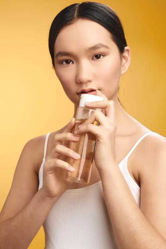 asian woman drinks from a clear bottle in a warm yellow studio, creating a calm skincare moment with natural, simple beauty and focused expression.