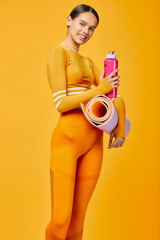Fitness woman in bright athletic wear stands against a yellow backdrop, holding a pink water bottle and rolled mat, ready for sport, workout, and healthy training.