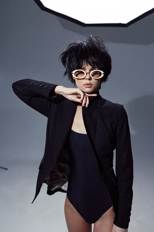 Stylish woman wearing white sunglasses and black blazer with confident expression in minimalist studio setting
