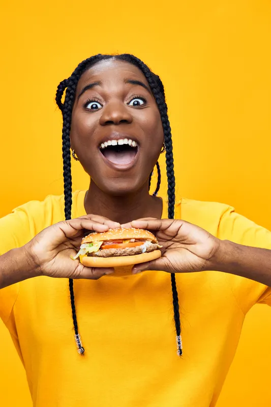 Joyful girl with braids holding a delicious burger against a vibrant yellow background, showcasing excitement and a love for food