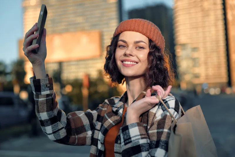 Woman selfie shopping urban smile candid lifestyle golden hour authenticity mindful living emotional storytelling as she poses with paper bags and phone, cozy beanie and plaid coat