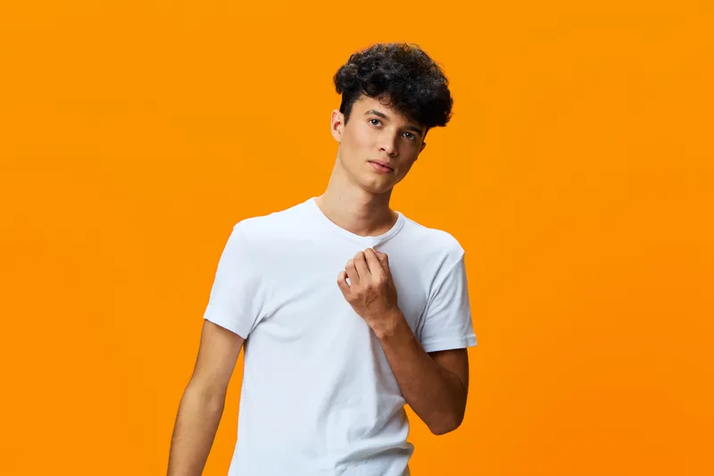 young man with curly hair wearing white t-shirt stands against bright orange background looking thoughtful and relaxed, contemporary casual style portrait.