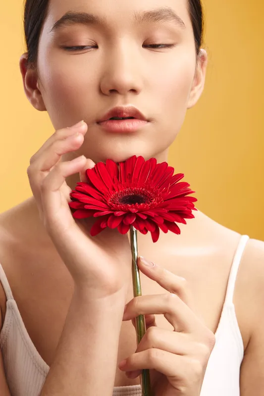 Asian woman with calm expression holds a red flower close to her face, eyes closed, creating a serene beauty shot with warm light and a soft yellow background.