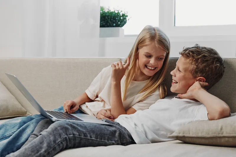 Happy children relax on sofa with laptop, smiling and enjoying digital entertainment together. Bright natural light fills cozy modern living room, concept of childhood, technology, and friendship.