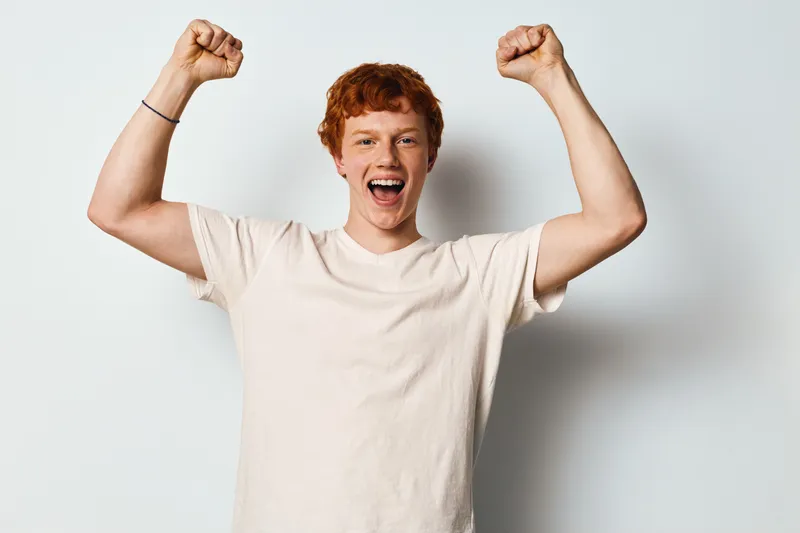 Young man with red hair smiling and raising fists in victory gesture on white background. Casual clothing, excited facial expression, positive energy and happy mood.