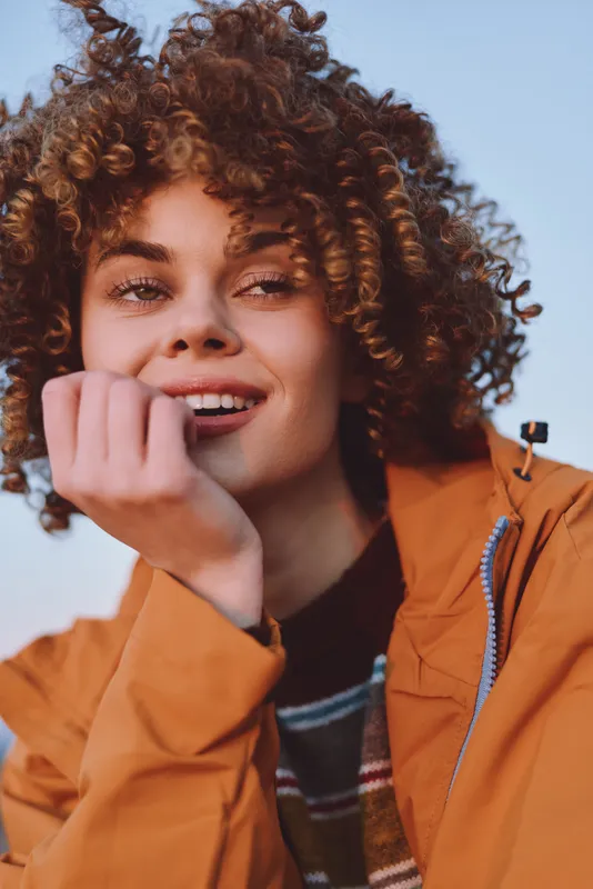 Woman lifestyle portrait outdoors, curly hair framing her face, smiling softly while touching her cheek, wearing an orange jacket, clear sky background, natural light, relaxed pose today