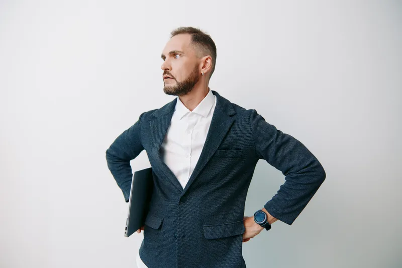 A professional man in a blazer stands with hands on hips, holding a laptop, against an isolated colored background. He projects confidence, focus, and determination, ready to lead and deliver impact.