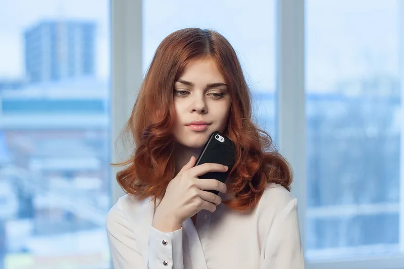 Рыжеволосая женщина в рубашке держит в руках телефон
