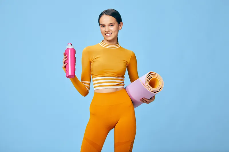 Woman in bright orange athletic outfit holds a pink water bottle and a rolled yoga mat against a blue backdrop, showcasing fitness, sport and wellness inspiration.
