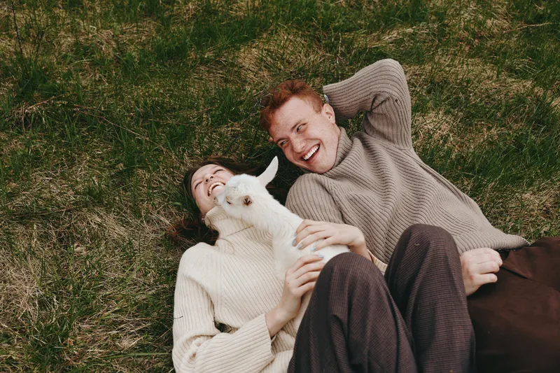 Joyful couple lying on grass with a playful goat kid, sharing a warm moment outdoors. Casual, cozy lifestyle scene highlighting connection, nature, and animal companionship.