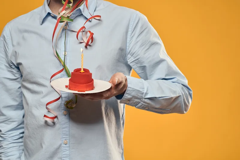 Мужчина в голубой рубашке держит красный торт со свечой на оранжевом фоне