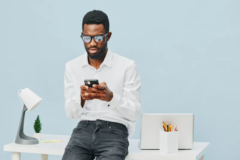 Professional young Afro-American man wearing white shirt and glasses, sitting at a modern office desk and looking at a smartphone screen, with a laptop and stationery in background, indoor studio shot