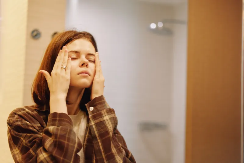 Woman with eyes closed and hands on temples, expressing stress and concentration in a casual plaid shirt. An intimate moment of relaxation and wellness in an indoor setting.
