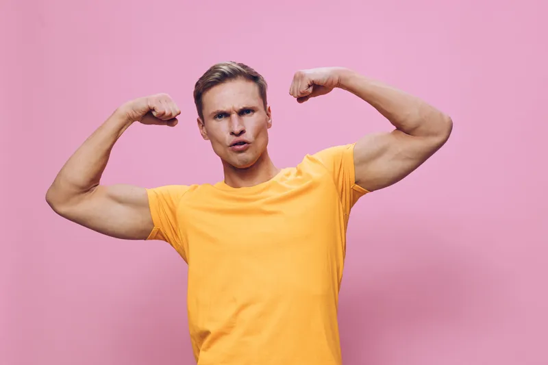 Confident young man with muscular arms wearing yellow t-shirt flexing biceps in studio against pink background. Portrait of strength, fitness, masculinity, healthy lifestyle, bodybuilder, confident po